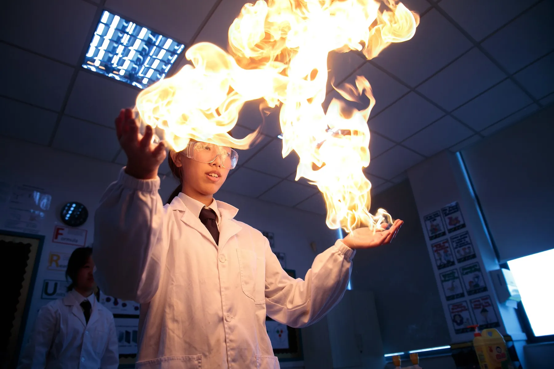A student in a lab coat and goggles holds a sheet of flame above her hands in a science demonstration