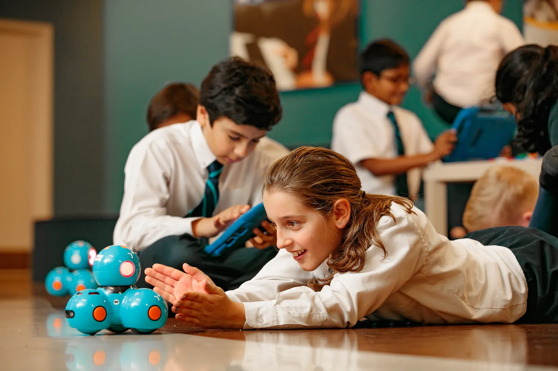 A delighted girl lies on the floor guiding a small coding robot as classmates work on tablets behind her