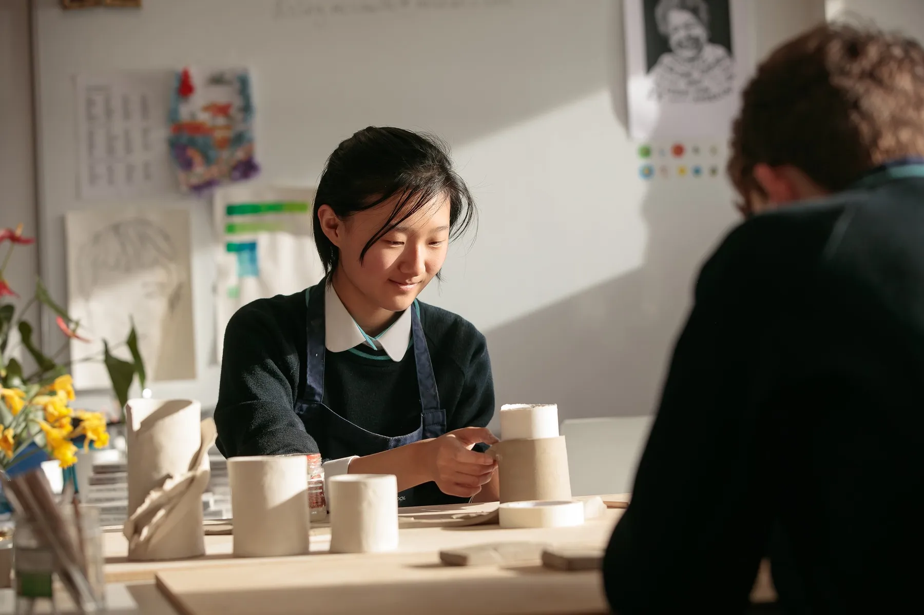 A student smiles quietly as she shapes a clay vessel in a sunlit art room