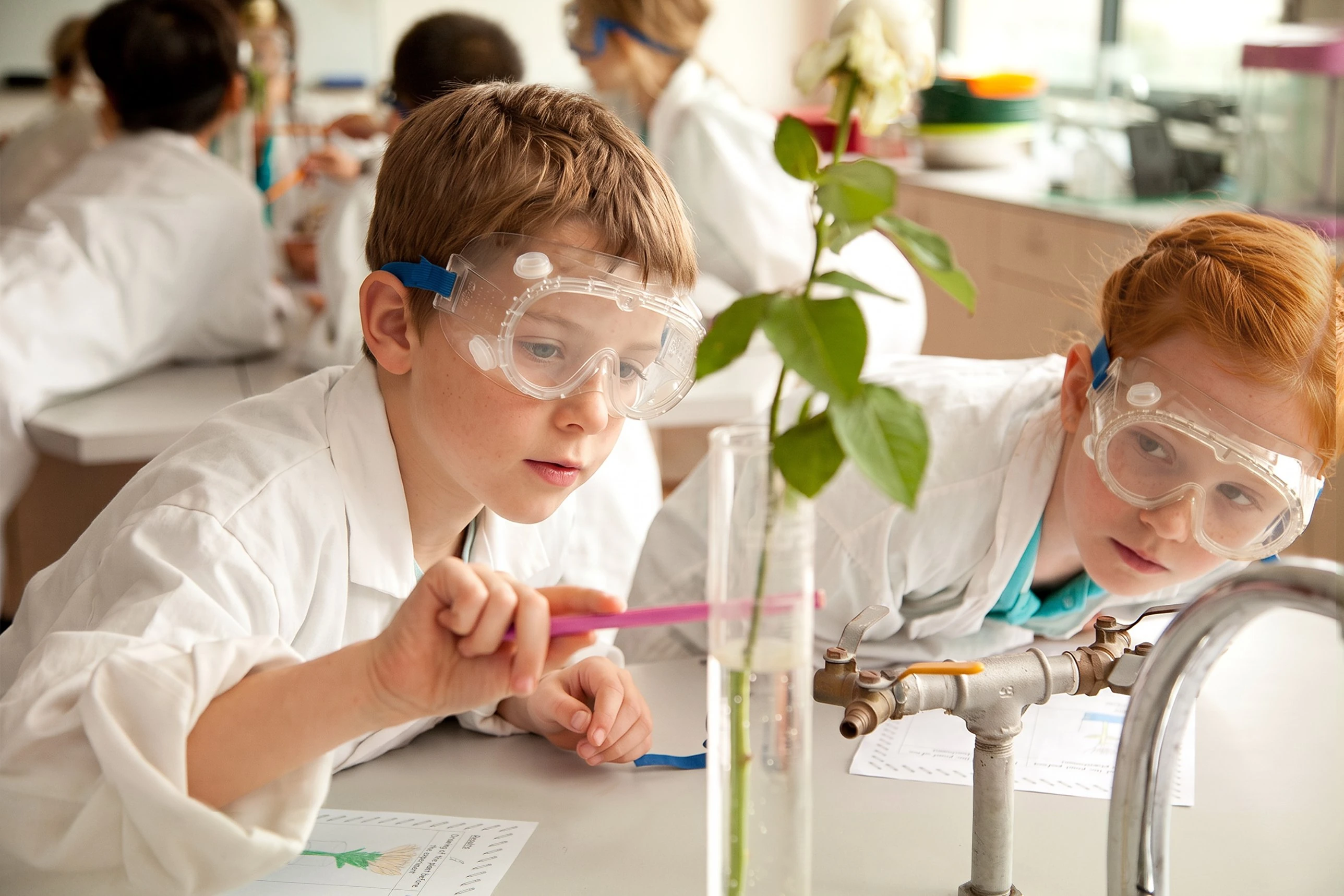 Two students in lab coats and safety goggles studying a plant experiment, framed in a camera viewfinder