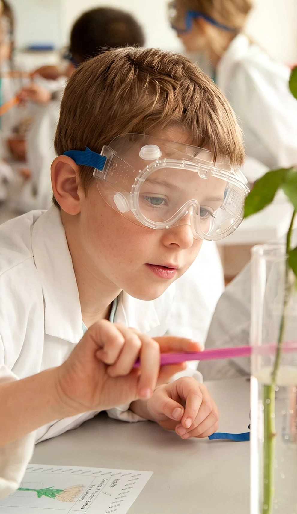 A student in a lab coat and safety goggles studying a plant experiment, framed in a camera viewfinder